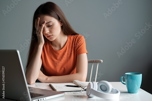 A young woman wearing an orange shirt looks overwhelmed while studying at a desk in her home. Generative AI.