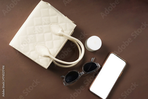 A cream-colored women's bag, sunglasses, a phone, and a moisturizer are placed against a brown background