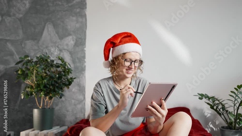 Woman in Santa hat sitting on bed with tablet and Christmas decorations around, enjoying cozy winter evening at home. Concept of holiday comfort and relaxation.