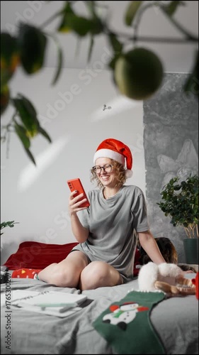 Relaxed woman in Santa hat and glasses lying on bed with smartphone and notebook, thinking and planning creative ideas during cozy winter evening.