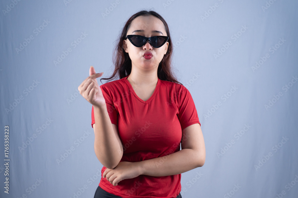 Fototapeta premium A woman in sunglasses and a red shirt makes a finger heart gesture with a pouting expression.