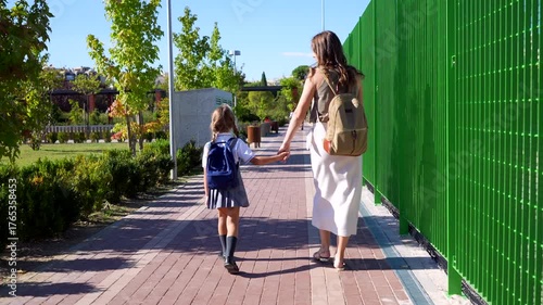 Young mother walking her little daughter wearing a uniform to school on a sunny day. A parent taking her child to elementary school, showing care, love, and support for her education