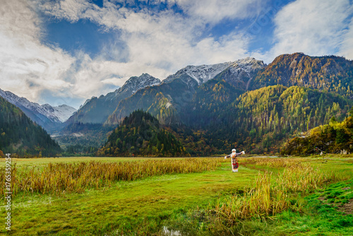 Traditional Tibetan woman standing in the wide green valley of Jiuzhaigou, China, surrounded by snow-capped mountains, autumn forest, and soft morning light under a blue sky.