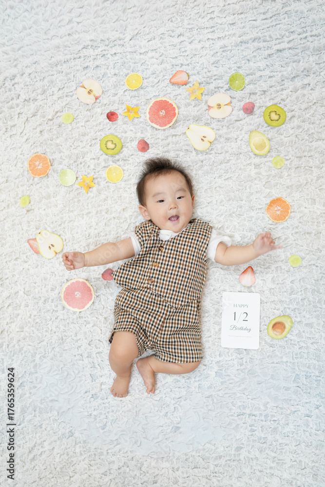 Naklejka premium A baby is laying on a white rug with a bunch of fruit around him. The fruit includes apples, oranges, and kiwis. The baby is wearing a brown and white outfit. The scene is playful and lighthearted