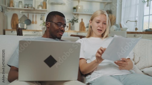 Man and woman discussing financial troubles at home, lady throwing papers in air . Portrait of young interracial spouses sitting on sofa in apartment and talking about utility bills and mortgage loan