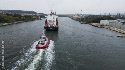 Aerial view of a red tugboat escorting a large cargo ship through the industrial port channel. Maritime navigation and harbor operations concept