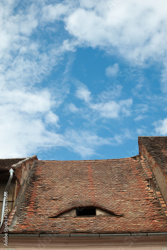  An old sloping roof with old tiles and a skylight, fragment of blue sky with clouds in the background.