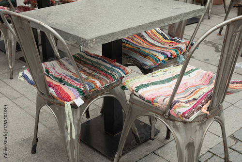 Metal chairs with colorful cushions and a table with a stone top, a café, a sidewalk.