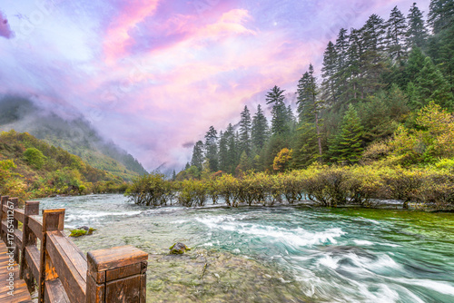 Misty forest morning along a wooden boardwalk beside a turquoise mountain river in Jiuzhaigou National Park, Sichuan, China. Tranquil autumn landscape under pastel pink sunrise skies.