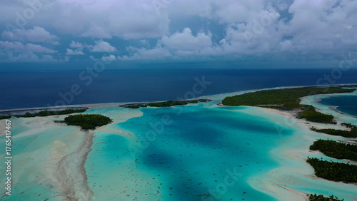Aerial view of the bright turquoise lagoon contrasting with the dark blue ocean, fringed by lush green islets under a cloudy sky, Rangiroa, Tuamotu Islands, French Polynesia.