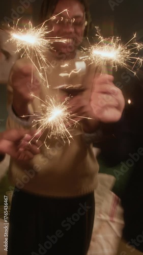 Vertical motion blur slowmo of diverse friends waving sparklers and raising glasses, celebrating Christmas together in warm home with glowing festive tree