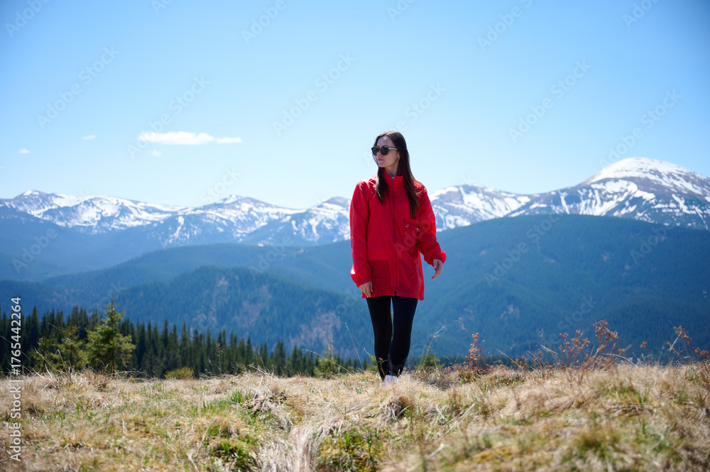 Naklejka premium Hiker girl on background of snowy mountains. Young female in red sport jacket and sunglasses hiking in the mountains. Incredible landscape reflects peace and freedom.