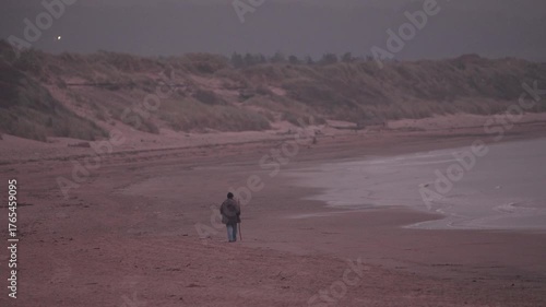 An elderly man walks alone along a quiet beach at sunset. Peaceful coastal evening scene evokes solitude, reflection, and the passing of time in nature