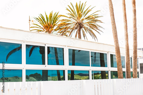 White modern building with large tinted windows reflects palm trees and distant beach, framed by crisp white fence. Harmonious facade merges architecture with tropical serenity inviting coastal living