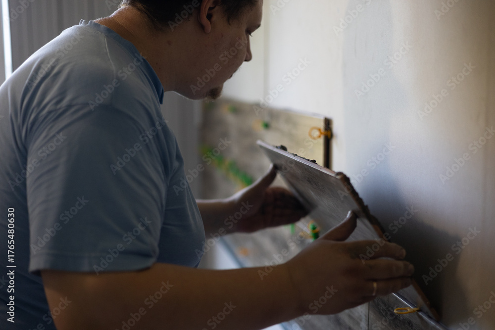 Obraz premium A man lays ceramic tiles on an interior wall while renovating or improving a home. The process of laying tiles symbolizes independent projects and skilled labor. Laying tiles on a wall in a house.