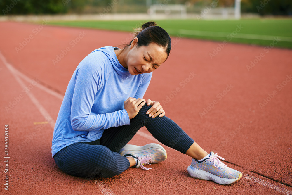 Fototapeta premium Female athlete holding painful knee sitting on outdoor stadium running track