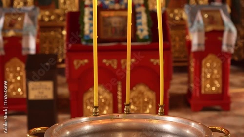 Three church candles burn softly near the baptismal font in an Orthodox temple. The golden light reflects on icons and creates a peaceful, sacred atmosphere of prayer and faith.