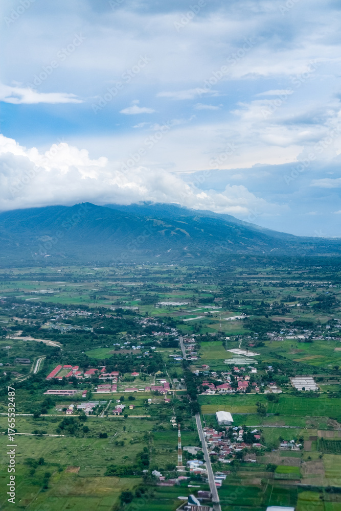 Fototapeta premium Majestic aerial view of a mountain valley surrounded by lush hills and clouds, overlooking a sprawling town below with green fields, scattered houses, and winding roads blending into the landscape.