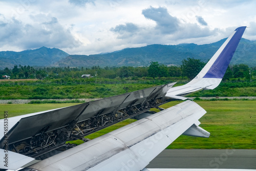 Aircraft wing with extended spoilers during landing rollout, showcasing aerodynamic braking in action as the plane touches down on the runway with green landscape and mountain views in the background.