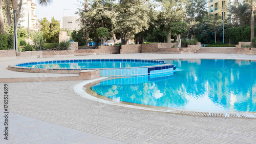 A serene swimming pool with clear blue water, surrounded by palm trees and landscaped gardens. The pool features a curved design and tiled edges.