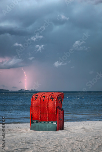 A red beach chair on a sandy beach of the Baltic Sea in the Kiel Fjord during a thunderstorm with a flash of lightning.