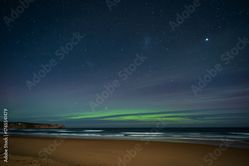 Quadro su tela The green southern lights of the Aurora Australis over the dark, blue ocean as waves break on the sandy beach