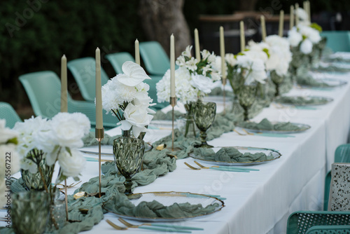 Decorated long table for a special occasion or event with vases with anthuriums, ivory taper candles, green goblets, gold cutlery and transparent plates, elegant outdoor wedding reception arrangement