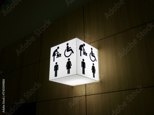 A glowing restroom sign showing icons for men, women, baby changing, and wheelchair access. The bright cube light contrasts with the dark modern interior wall.