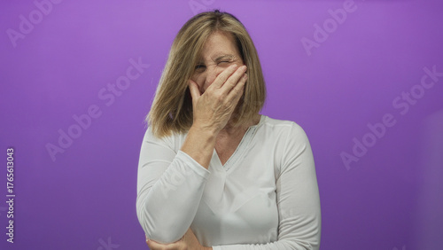 Foto Middle-aged woman wearing white blouse covers mouth with hand on purple backdrop in studio; discomfort pain distress toothache