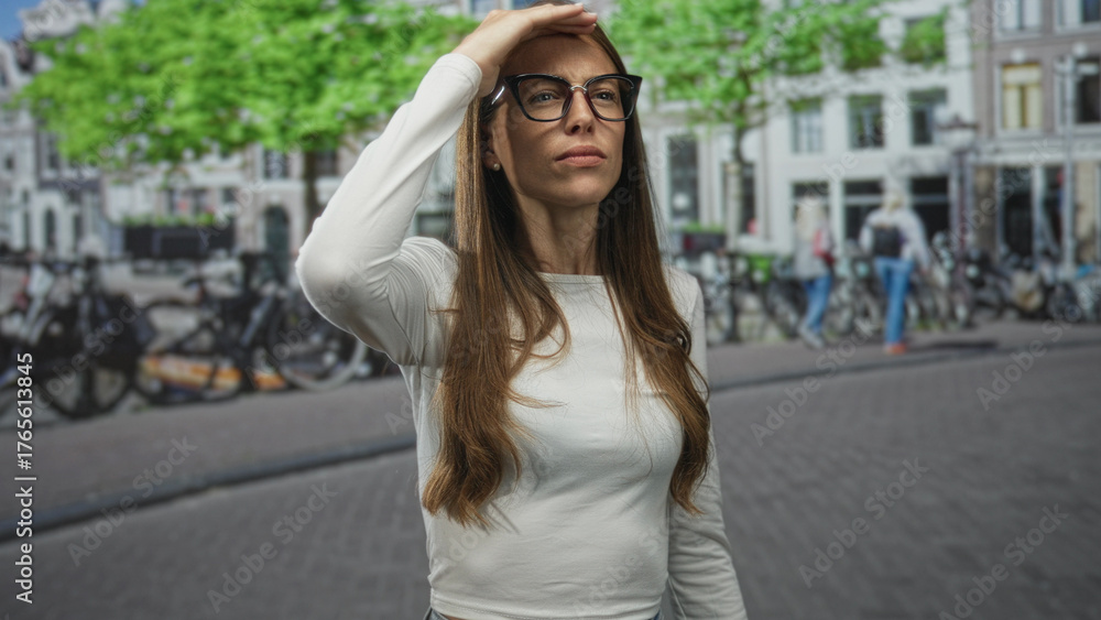 Fototapeta premium Young woman with glasses shielding eyes with hand to forehead while squinting on street in city; searching.