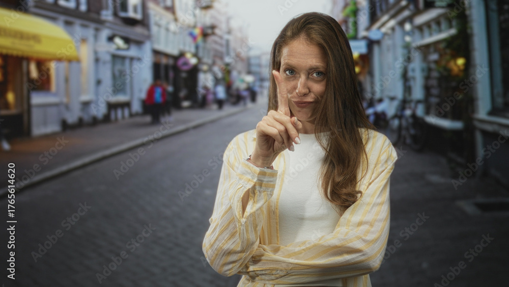 Fototapeta premium Woman with index finger raised and crossed arms on a cobblestone street in a striped shirt; playful warning.