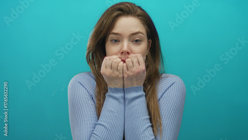 Young caucasian woman with long brown hair holds clenched fists to mouth in studio setting with turquoise backdrop; anxiety.