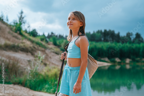 A young girl with a backpack gazes upward, smiling slightly under a cloudy sky, with lush greenery and a serene lake in the background.