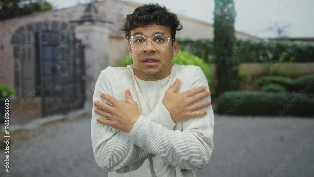 Fototapeta premium Young man hugs bare shoulders outside stone building entrance as he shivers in light sweater; anxiety.