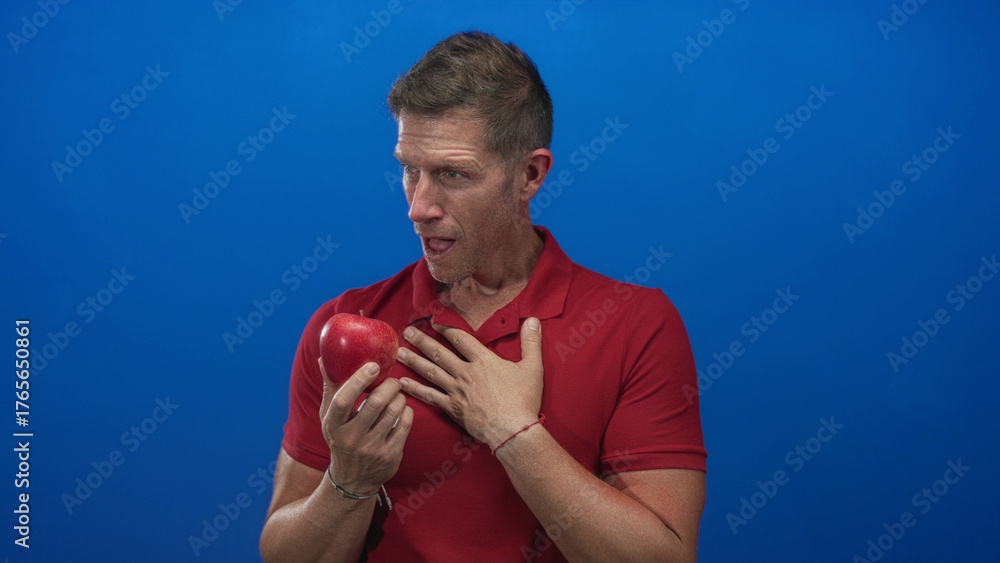 Φωτογραφία Man wearing red polo shirt holding red apple in one hand and resting other hand on chest with slight smile in blue studio; temptation