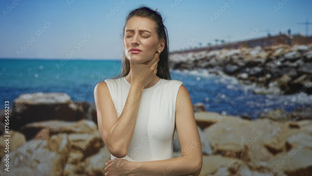 Φωτογραφία Woman with hand on neck showing neck pain in a seaside studio set with rocky coast and turquoise sea under clear sky; pain
