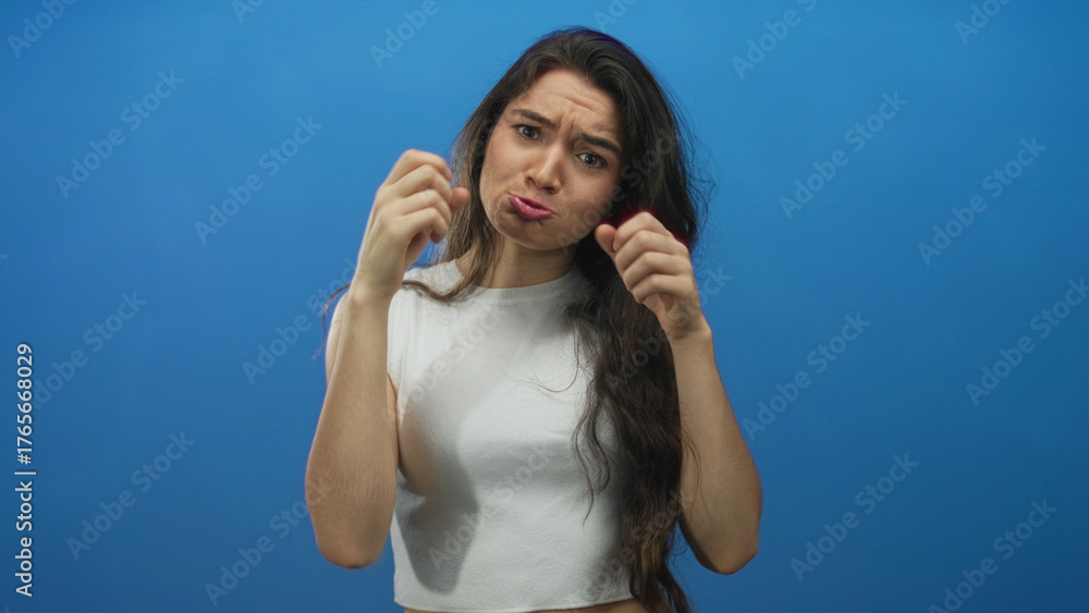 Fototapeta premium Young hispanic woman mimics crying gesture with fists to cheeks in blue studio; sadness vulnerability empathy.