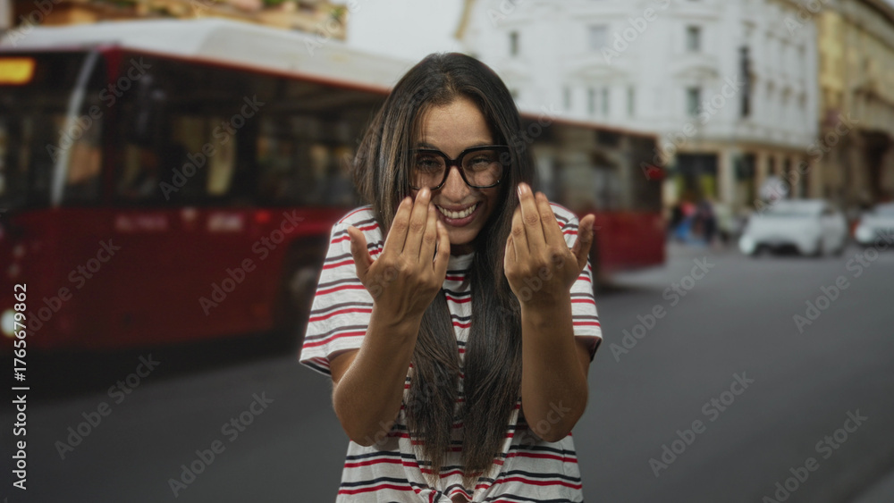 Fototapeta premium Woman with glasses and striped shirt beckons with hands on a busy city street beside a red bus and classical building; playful.
