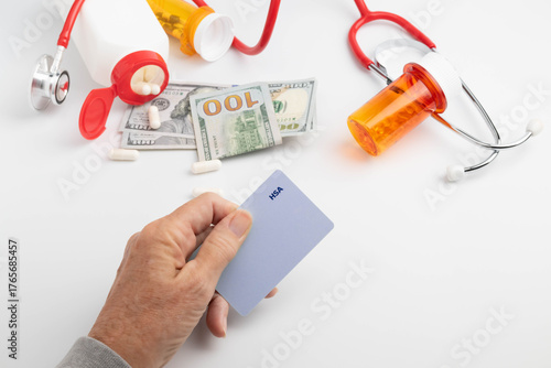 High cost of healthcare concept.  Hand holding a purple HSA card across white background with pill bottles, medicine, stethoscope, and American currency, one-hundred-dollar bills.