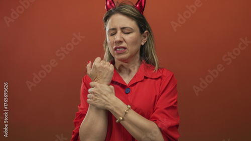 Foto Woman clutching wrist with pained expression wearing devil horns headband and red shirt in red studio; discomfort
