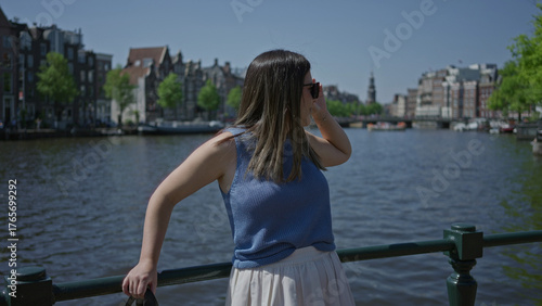 Photography Woman enjoying sunlight by amsterdam canals with historic architecture in the background, wearing sunglasses, capturing a relaxed moment in the urban outdoors of the netherlands