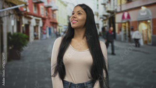 Young latin woman smiling while looking upward with head tilted on sunlit city street under soft natural light; optimism. Free Download Young latin woman smiling while looking upward with head tilted on sunlit city street under soft natural light; optimism. - GFXLoad