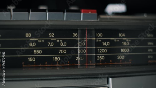 Closeup of a vintage radio tuner scale with a red needle moving left across frequency bands. The analog display shows a search for stations on an old silver boombox receiver with buttons.