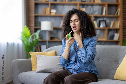 Woman on couch at home treating sore throat with medicated throat spray, appearing unwell and uncomfortable while seeking relief from cold or flu symptoms indoors