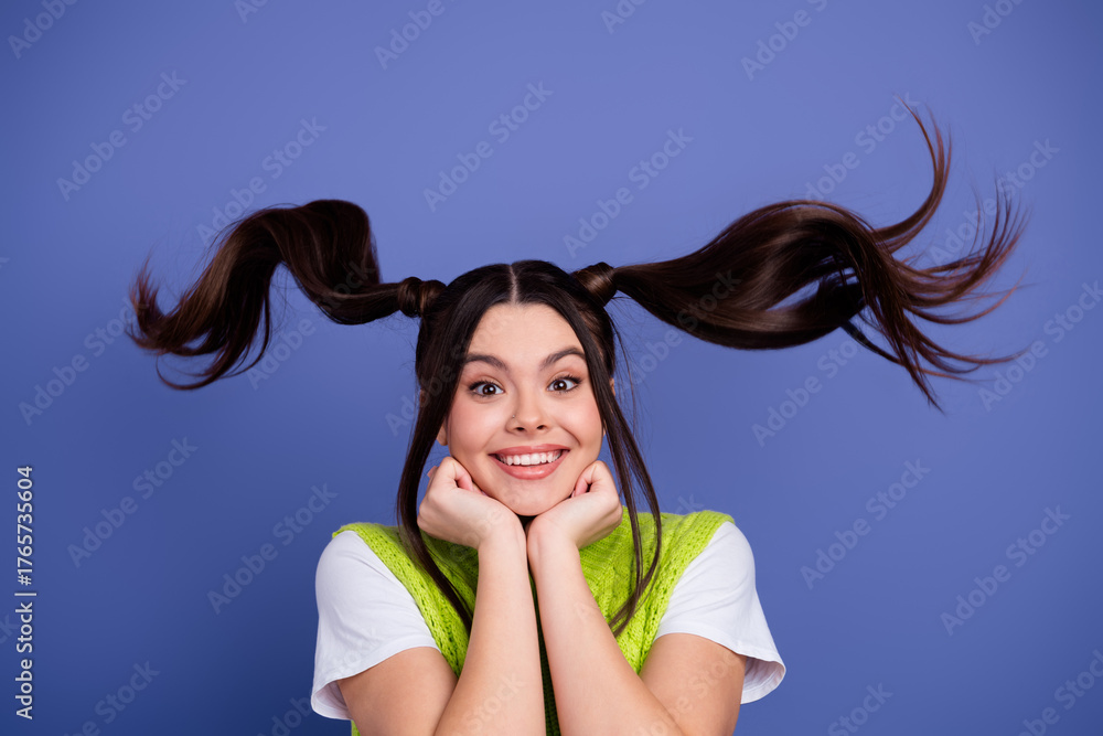 Naklejka premium Joyful young woman with playful pigtails wearing a lime green vest smiles with hands under chin against a violet blue background
