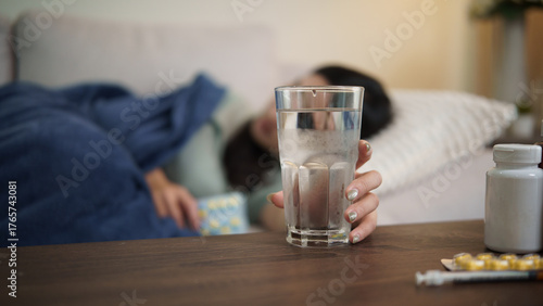 Sick woman lying on sofa reaching for a glass of water, feeling weak and tired while recovering from illness at home.