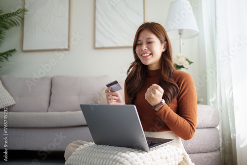 Excited young woman holding credit card and celebrating success while using laptop at home. Concept of online shopping and digital payment.
