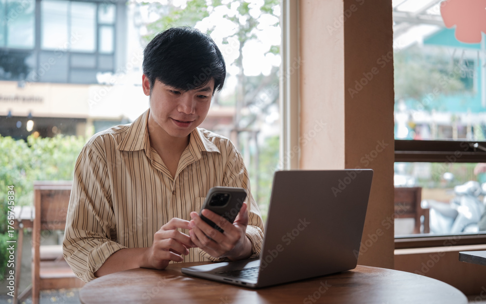Fototapeta premium Smiling young man interacting with his mobile phone while working on laptop in a modern cafe.