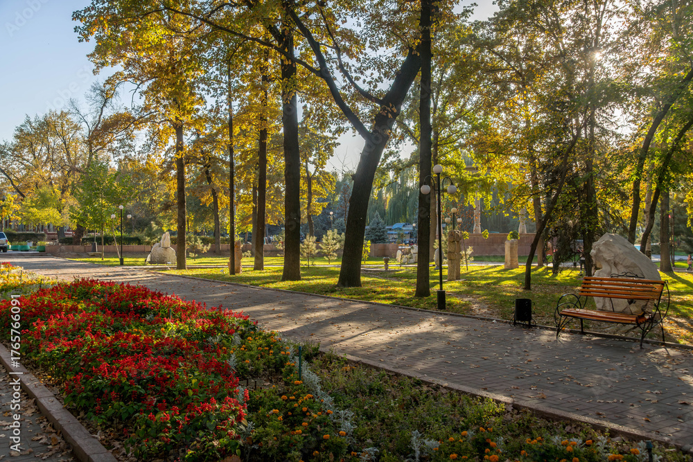 Naklejka premium Flowers in park at Bishkek Oak Park with tall autumn trees, and seasonal garden colors.