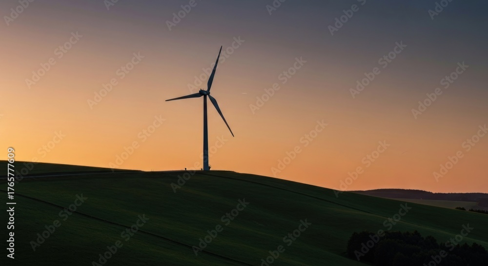 Fototapeta premium Wind turbine silhouetted atop a grassy hill against a warm sunset sky